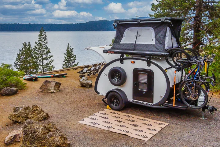 An RV rental teardrop trailer with a rooftop tent and bikes is parked at a scenic lake campsite with paddleboards.. Bean Trailer Bean Stock 2022
