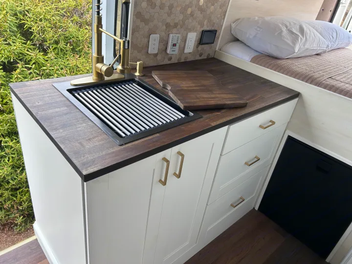 Elegant RV rental features a compact kitchen with gold faucet, dark wood counter, white cabinets, & bed. Ideal for mobile comfort.. Ram Promaster 2500 2022