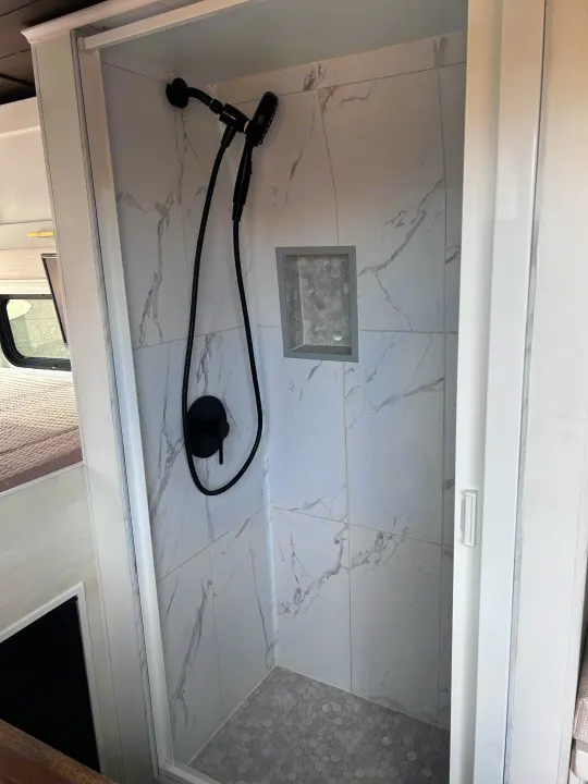 Compact RV rental shower with white marble-look tiles, hexagonal gray floor, and sleek black fixtures.. Ram Promaster 2500 2022