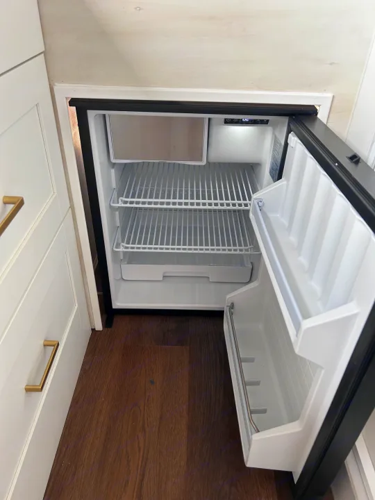 An open, empty mini-fridge in an RV rental, featuring wire shelves and a compact freezer, set within white cabinetry.. Ram Promaster 2500 2022