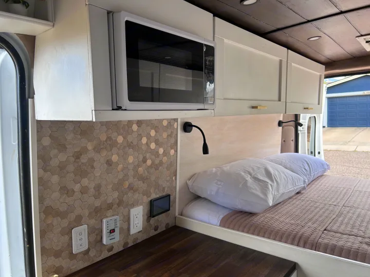 Sleek RV rental interior showcases a microwave, overhead cabinets, hexagon tile backsplash, and a cozy bed area.. Ram Promaster 2500 2022