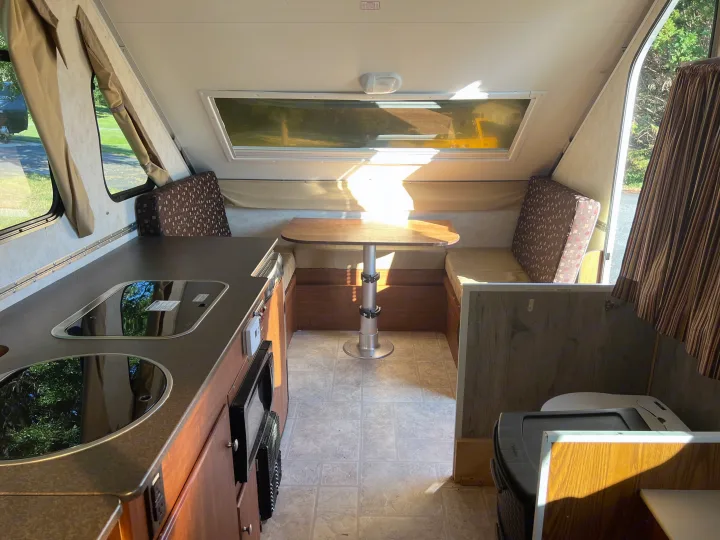 Inside an RV rental: Compact kitchen with a cooktop/sink, dining booth, and sunlit windows. A functional & cozy camper interior.. A Liner A Liner Popup Trailer 2013