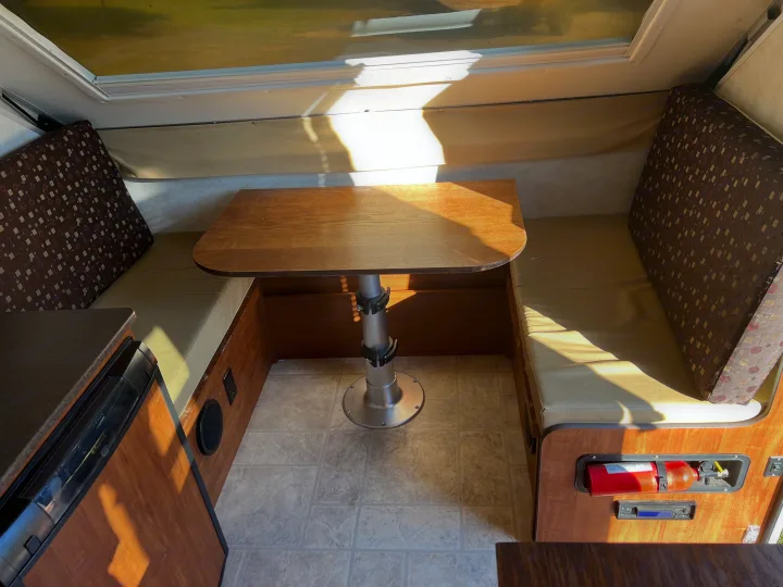 Interior of an RV rental: a cozy dinette with a wooden table, comfortable bench seating, compact fridge, and fire extinguisher.. A Liner A Liner Popup Trailer 2013