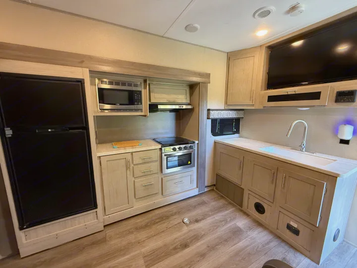 Modern RV rental kitchen with fridge, microwave, stove, oven, sink, and TV, featuring light wood cabinets.. Coachmen Catalina 2022