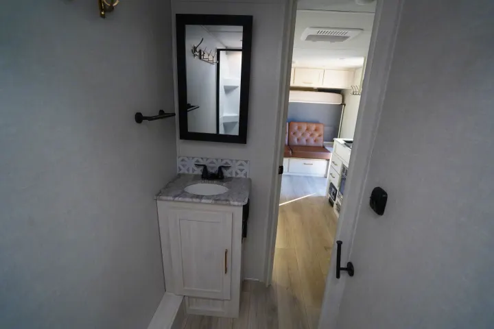 Inside an RV rental: Bathroom with mirror & vanity, leading to a living area with a brown bench and light floors.. Forest River Surveyor Legend 2024
