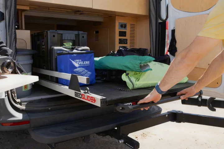 RV rental van: A person extends a Bedslide cargo tray, revealing organized gear & leveling blocks. Efficient storage for adventures.. Mercedes-Benz Sprinter 2020