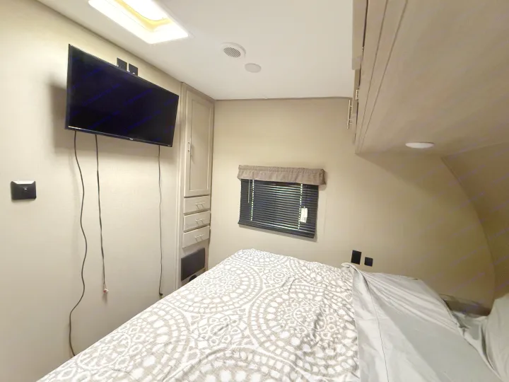Inside an RV rental bedroom: wall-mounted TV, comfy bed with a patterned duvet, tall storage unit, and a window with blinds.. Coachmen Catalina 2022