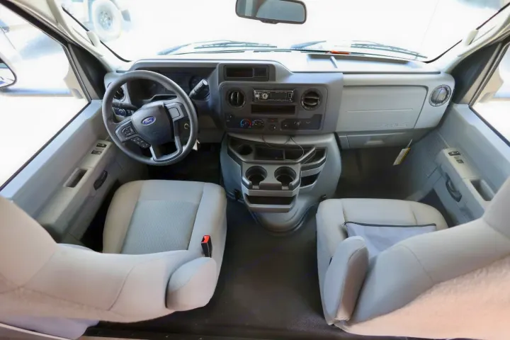 RV rental's Ford E-Series front cabin, displaying the steering wheel, dashboard with radio, and light grey driver/passenger seats.. Coachmen Freelander 2023