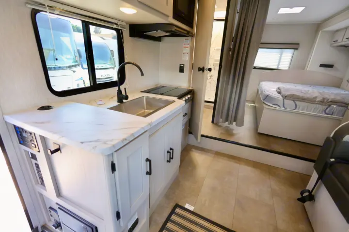 Interior of an RV rental: a compact kitchen with a sink and stove, leading to a bedroom and visible bathroom area.. Coachmen Freelander 2023
