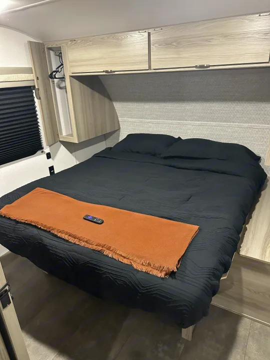 Modern RV rental bedroom with a black bed, rust throw, remote, overhead storage, and pleated blinds.. Forest River Salem Cruise Lite 2024