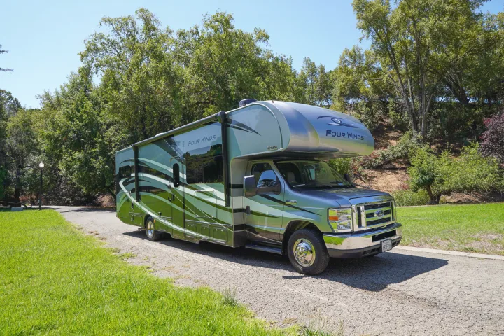A modern green and silver Four Winds rv rental parked on a sunny road, surrounded by lush trees and grass.. Thor Four Winds 2020