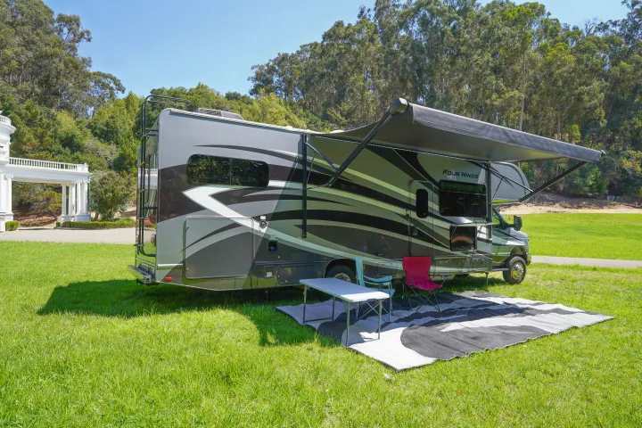 An RV rental parked on a grassy field, featuring an extended awning, chairs, table, and mat for outdoor relaxation.. Thor Four Winds 2020
