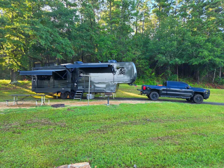 Dark grey fifth-wheel RV rental and truck parked at a serene, green campsite surrounded by trees.. Keystone RV Cougar 2024