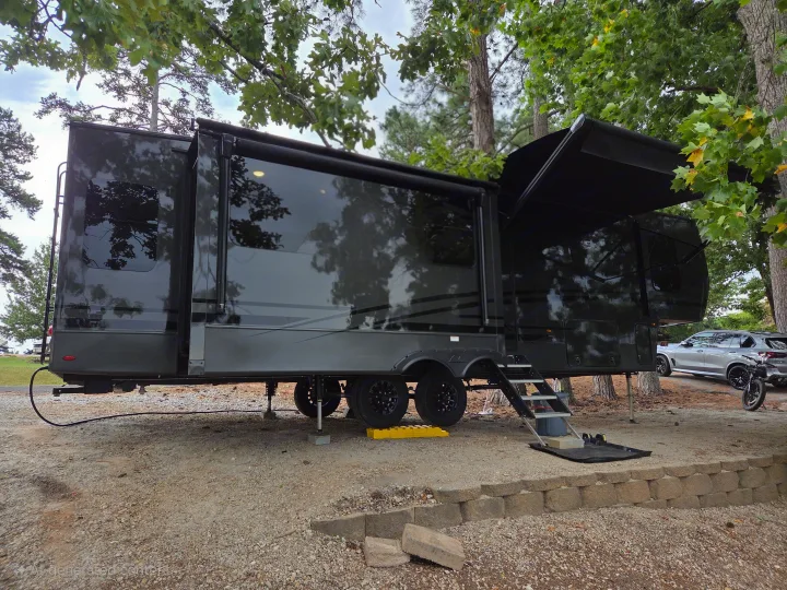 Modern dark fifth-wheel RV rental set up at a wooded campsite, with an SUV and electric bike parked nearby.. Keystone RV Cougar 2024