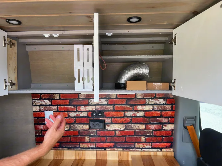 Inside this rv rental, open overhead cabinets reveal storage. A brick backsplash with outlets sits above a wooden countertop.. Mercedes Sprinter 2011