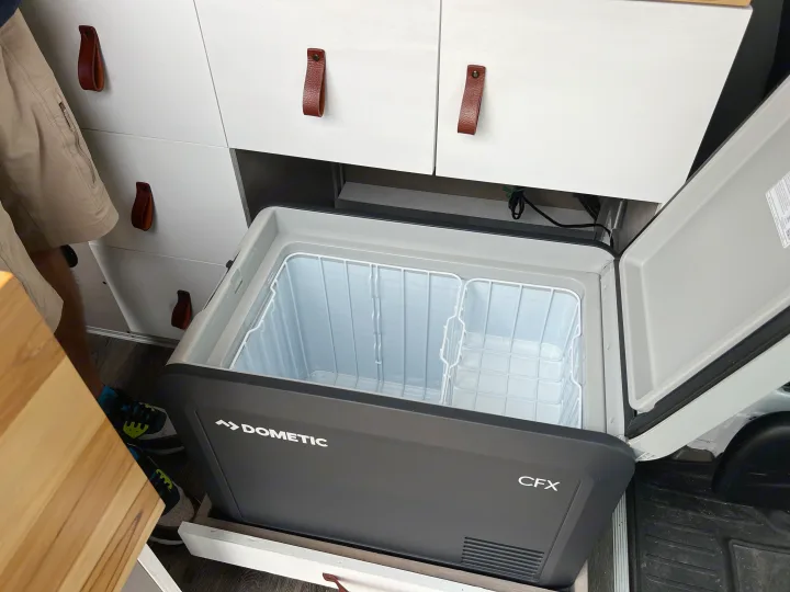 Open Dometic CFX portable fridge in an rv rental camper van, showing wire baskets & white cabinets, ready for travel.. Mercedes Sprinter 2011