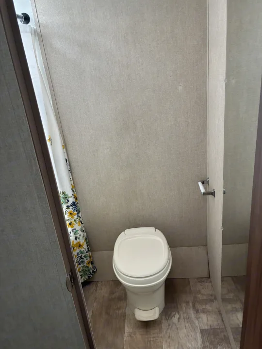 Inside an RV rental: a compact bathroom featuring a white toilet, floral shower curtain, and wood-look floor.. Gulf Stream Ameri-Lite 2019