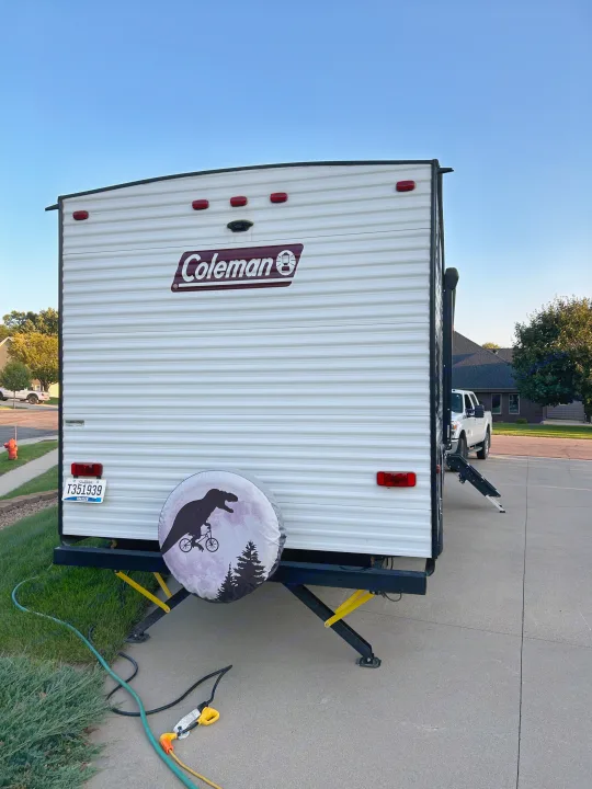 Rear of a Coleman RV rental parked in a driveway, featuring a distinctive T-Rex on bicycle spare tire cover.. Dutchmen Coleman Lantern 2020