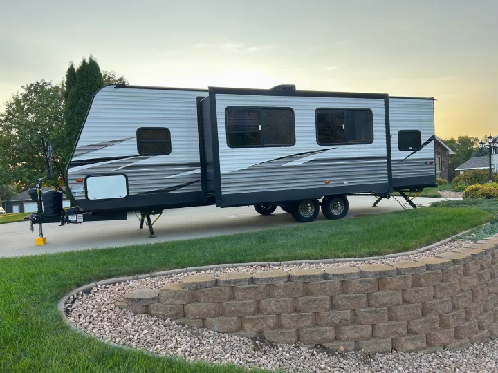Grey RV rental travel trailer with an extended slide-out, parked on a residential driveway near a stone wall.. Dutchmen Coleman Lantern 2020