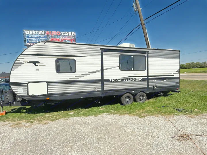A white and grey Trail Runner travel trailer (RV), ideal for rental, is parked on grass. An auto care sign is in the background.. Heartland Trail Runner 2022