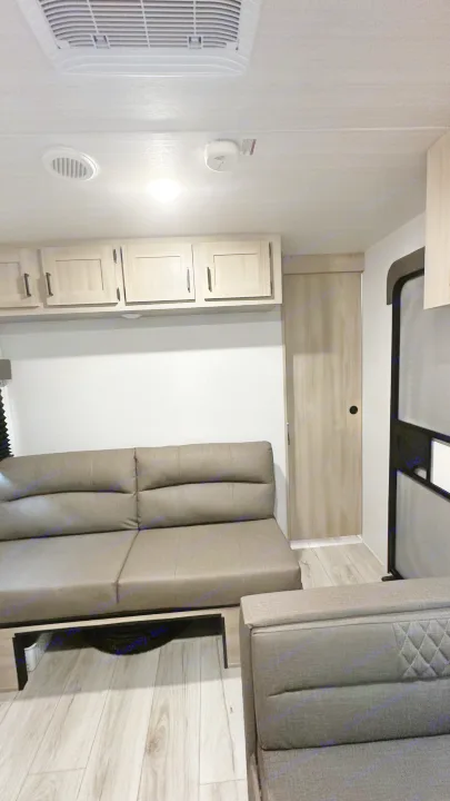RV rental living area featuring a long gray sofa, light wood overhead cabinets, and light plank flooring. Includes AC unit and door.. East to West Silver Lake 2024