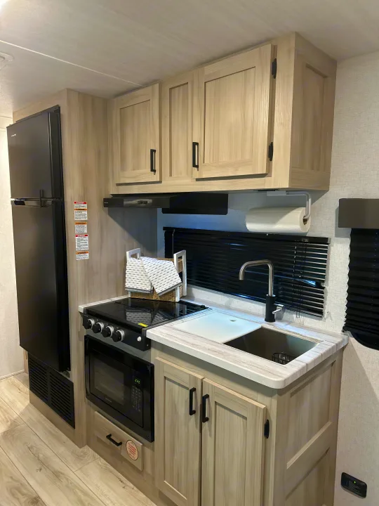 Bright rv rental kitchen with light wood cabinets, black refrigerator, stove, microwave, and stainless sink area.. East to West Silver Lake 2024