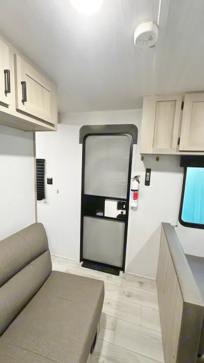 Inside an RV rental: light cabinets, wood-look floor, sofa, and entry door. Fire extinguisher and smoke detector visible.. East to West Silver Lake 2024