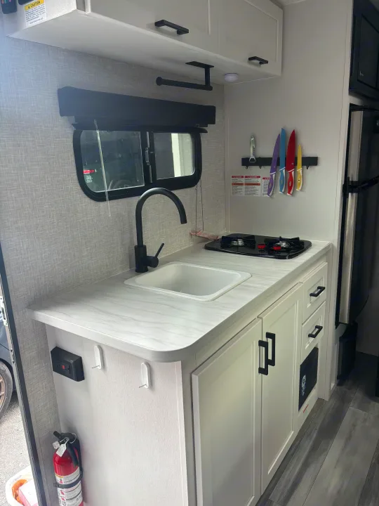 Explore this modern RV rental kitchen: sleek sink, two-burner stove, white cabinets, and colorful knife storage.. Jayco Jay Feather Air 2025