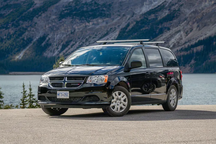 Black camper van RV rental (Dodge Grand Caravan) by a Canadian lake, mountains in background. Ideal for scenic travel.. Dodge Converted Dodge Caravan 2020