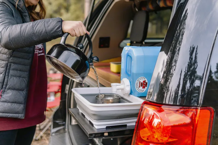 rv rental: Woman pours kettle water into a collapsible basin next to a blue jug, showcasing a convenient camper kitchen setup.. Dodge Converted Dodge Caravan 2020