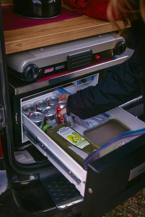 RV rental camping kitchen: Pull-out fridge drawer full of cold drinks and food below a Coleman propane stove for easy meals.. Dodge Converted Dodge Caravan 2020