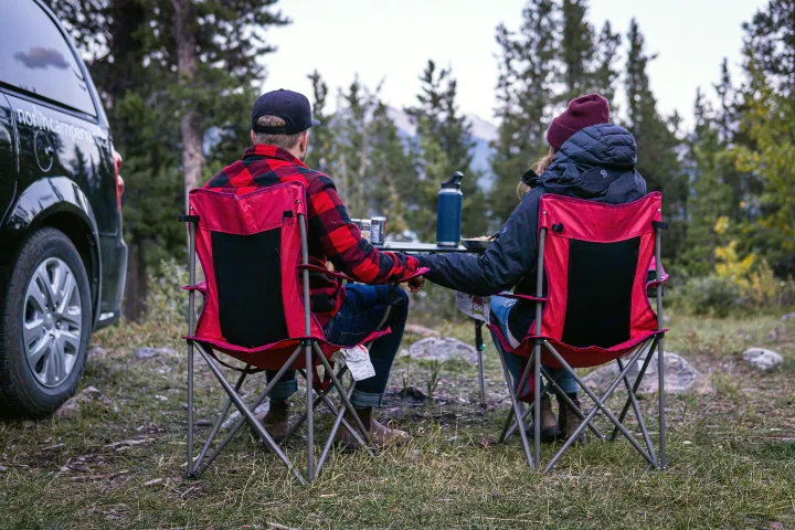 From behind, a couple in camping chairs holds hands, enjoying mountain & forest views from their RV rental.. Dodge Converted Dodge Caravan 2020