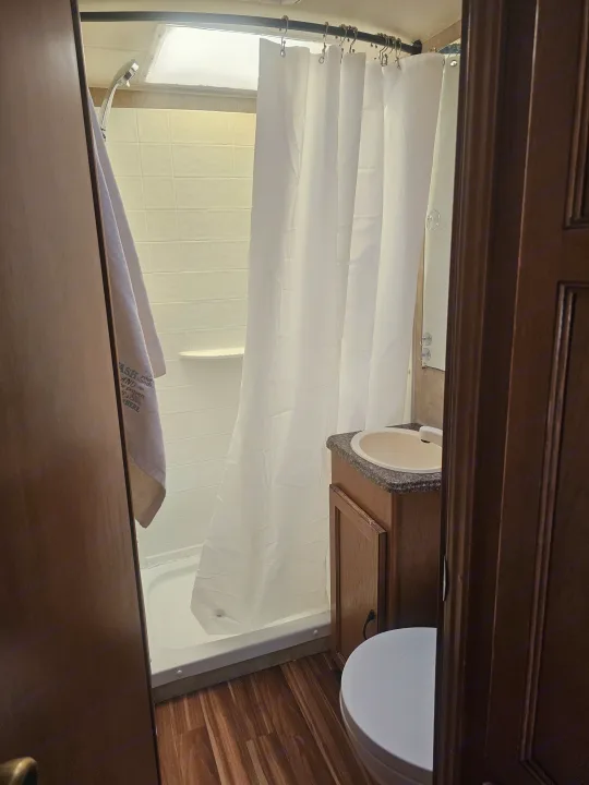 Compact RV rental bathroom with a white shower curtain, visible toilet, and sink vanity. Features wood-look flooring.. Coachmen Freelander 2014
