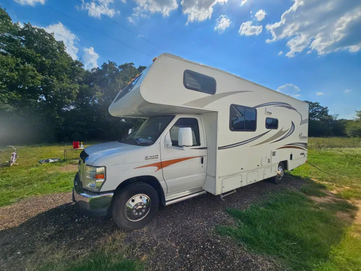 This white Class C rv rental is parked on a gravel spot amidst lush green grass and trees, under a bright, sunny sky.. Coachmen Freelander 2014