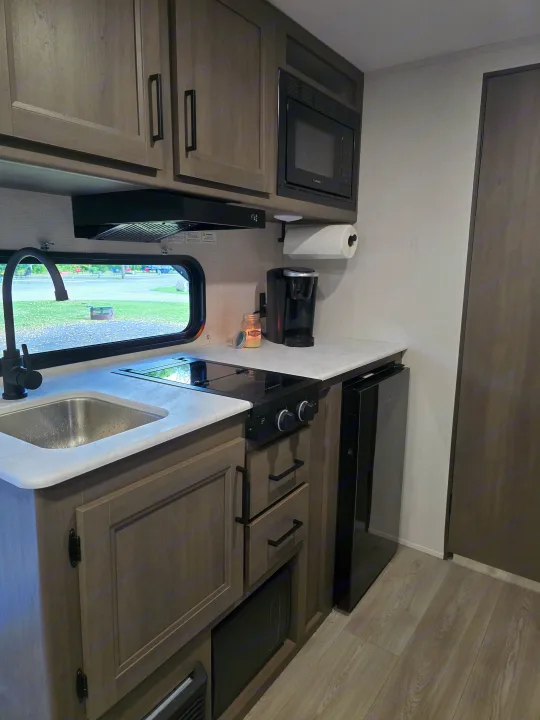 Compact RV rental kitchen: sink, stovetop, microwave, coffee maker, fridge, and a peaceful campground view.. Grand Design 151BH 2025
