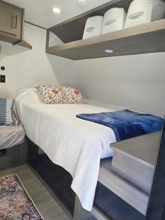 Cozy RV rental bedroom featuring a compact bed with white bedding, decorative pillows, and practical built-in overhead storage shelves.. Grand Design 151BH 2025