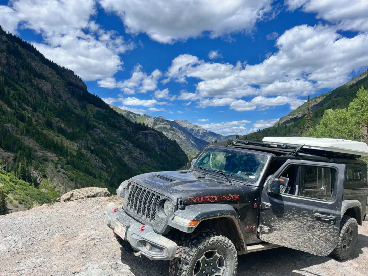 Mud-splattered Jeep Mojave with rooftop tent on a mountain trail, ideal for an RV rental adventure.. Custom Jeep Gladiator Mojave 2021
