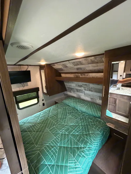 Step into comfort! This RV rental bedroom features a green patterned bed, wall-mounted TV, window, and mirrored storage.. Dutchmen Coleman Lantern 2020