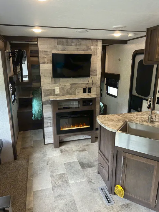 Modern RV rental living space featuring a kitchen island with sink, electric fireplace, TV, and bedroom beyond.. Dutchmen Coleman Lantern 2020