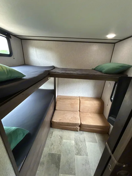 RV rental bunkhouse showing two beds with green pillows, plus a lower convertible cushion bed, all on gray flooring.. Dutchmen Coleman Lantern 2020