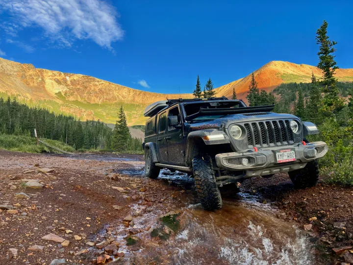 A black Jeep Gladiator with rooftop tent tackles a rocky mountain trail and stream. An epic scene for your next RV rental adventure!. Custom Jeep Gladiator Mojave 2021
