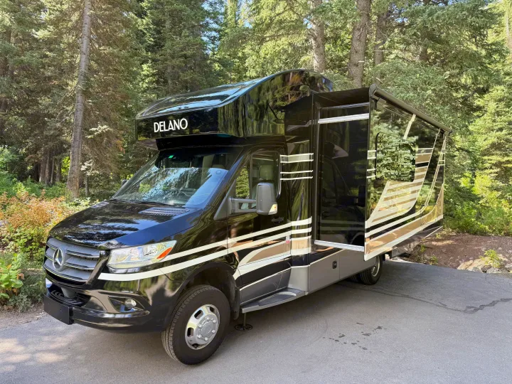 Black DELANO RV rental, a Mercedes-Benz Sprinter motorhome, parked amidst green trees with its slide-out extended.. Thor Delano 2022