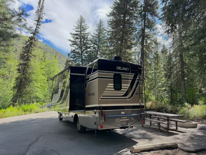 This grey and black RV rental is parked at a forested mountain campsite, ready for adventure with a picnic table nearby.. Thor Delano 2022