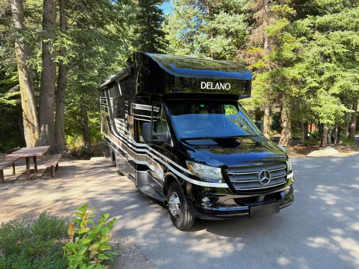 Stylish black Mercedes-Benz Delano Class C RV rental parked at a serene campsite with evergreen trees and a picnic table.. Thor Delano 2022