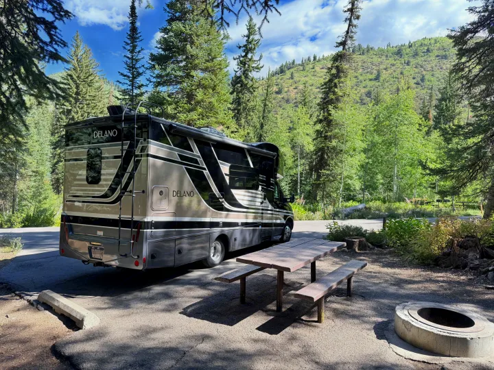 A 'DELANO' RV rental is parked at a scenic campsite with a wooden picnic table and fire pit, surrounded by tall trees and mountains.. Thor Delano 2022