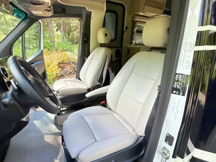 Inside a spacious RV rental: light-colored driver and passenger seats, cockpit, and nature views.. Thor Delano 2022