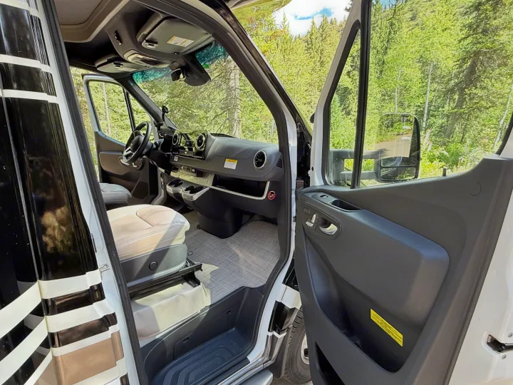 RV rental: Open door of a white camper van showing its modern interior, dashboard, and a lush green forest outside.. Thor Delano 2022