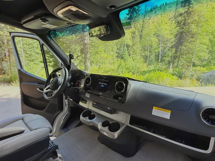 Inside a Mercedes-Benz RV rental camper van, showing the dashboard, steering wheel, and a scenic forest view through the windshield.. Thor Delano 2022