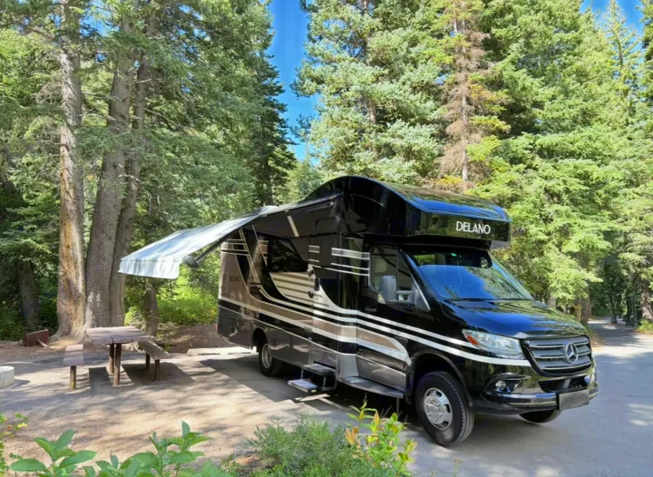 Black Delano RV rental with extended awning parked in a sunny, tree-filled campground next to a picnic table.. Thor Delano 2022