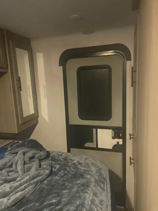 RV rental interior: A bed with a gray blanket, a mirrored cabinet on the left, and a main door marked "EXIT" on the right.. Dutchmen Coleman Light 2022